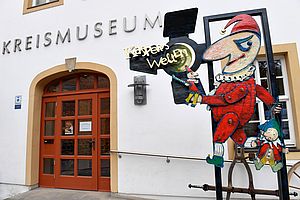 Es ist die Eingangstür vom Kreismuseum Bad Liebenwerda zu sehen. Rechts steht eine übermannsgroße Kasperfigur, die den Schriftzug Kaspers Welt in der Hand hält. 