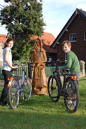 Holzskulptur - Mönch mit Fisch - in Fischwasser, davor zwei Kinder mit Fahrrad