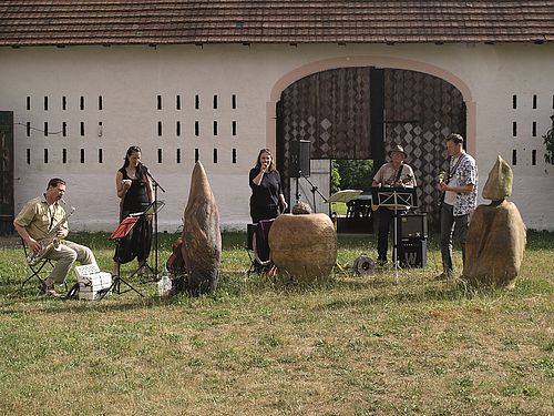 Vor einer Scheune mit einem großen Tor spielt eine Musikgruppe mit fünf Personen. Davor stehen drei Kunstskulpturen