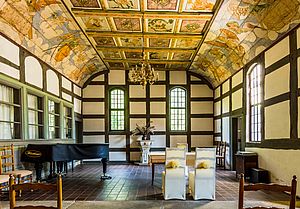 Innenraum einer kleinen Kirche mit Fachwerk, auf der linken Seite steht ein Flügel an der hinteren Wand zwischen den Fenstern ein Taufstein. Darauf steht ein Blumenstrauß. Im Vordergrund stehen zwei weiß bezogene Stühle mit der Rückseite zum Betrachter und davor ein Tisch