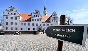 Im Hintergrund ist das Schloss Doberlug zu sehen. Im Vordergrund steht ein Wanderwegweiser mit dem Schriftzug `erlebnisREICH Naturpark`
