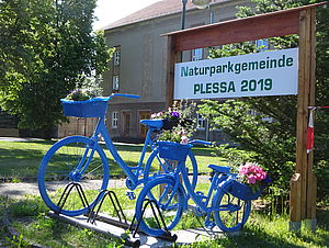 vor dem Kulturhaus Plessa stehen zwei blaue Fahrräder zur Deko mit Blumen dahinter ist ein Schild "Naturparkgemeinde Plessa 2019"