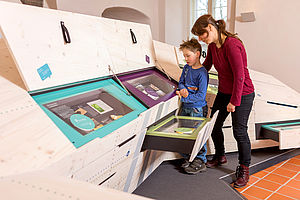 zwei Teile des Ausstellungskörpers `erlebnisREICH Naturpark`sind aufgeklappt, eine Schublade aufgezogen. Eine Frau und ein Junge schauen sich die aufgeklappten Tafeln an.