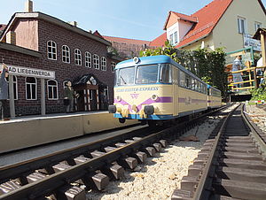 Ein frontaler Blick auf eine fahrende Modelleisenbahn kurz vor einem Modellbahnsteig. Im Hintergrund ist das Gebäude des Natoureums erkennbar. 
